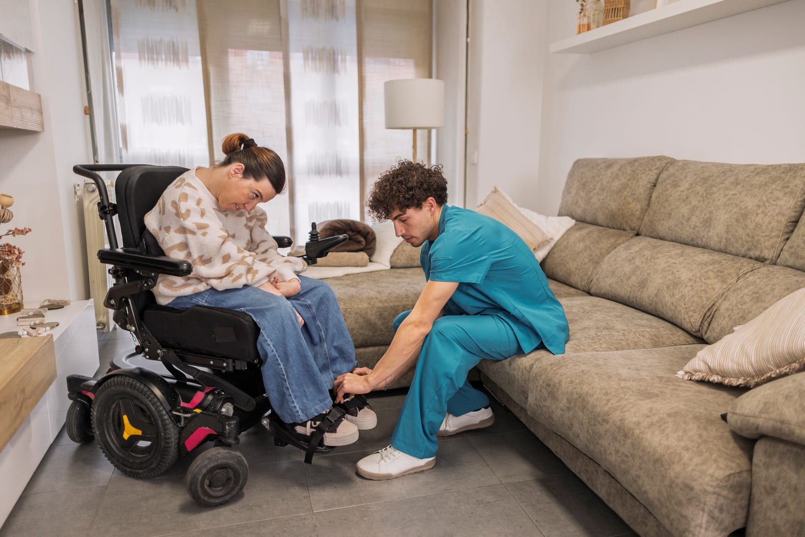 A caregiver kneeling to help a young woman using a wheelchair settle in at home